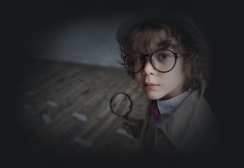 Colour photograph of small boy holding a magnifying glass, wearing a hat, large glasses, bow tie and trench coat.