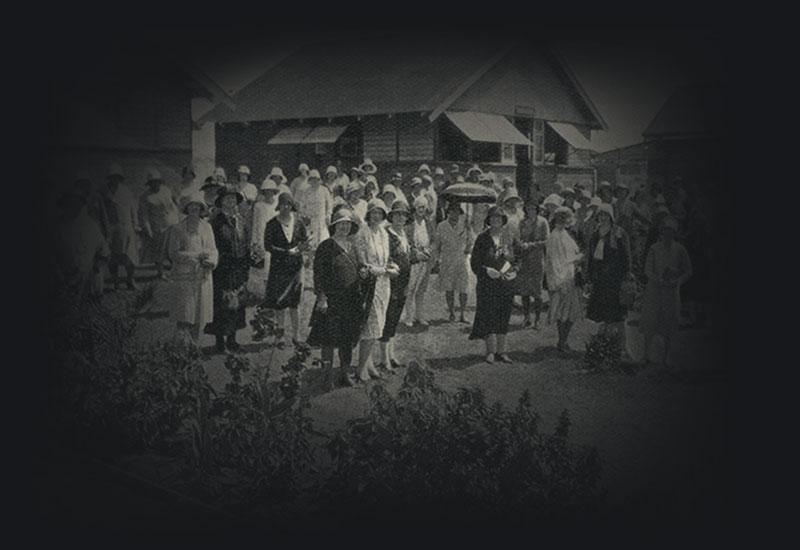 Women wearing hats – standing together on a lawn amongst large timber huts.