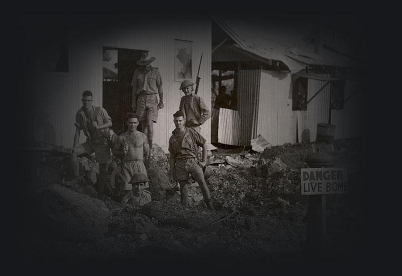Bomb disposal squad of 6 men standing around a bomb crater, near a shed and a danger live bomb sign.