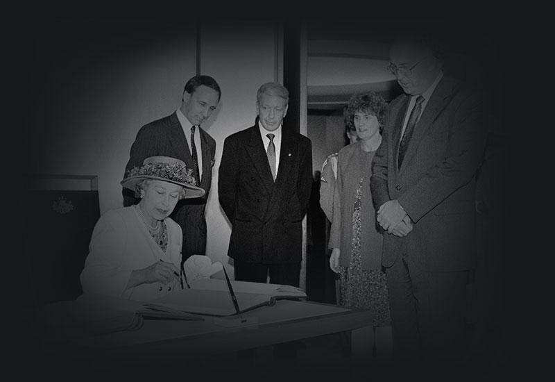 The Queen sitting at a desk, signing a document, in Parliament House. Prime Minister Paul Keating is in the background. 