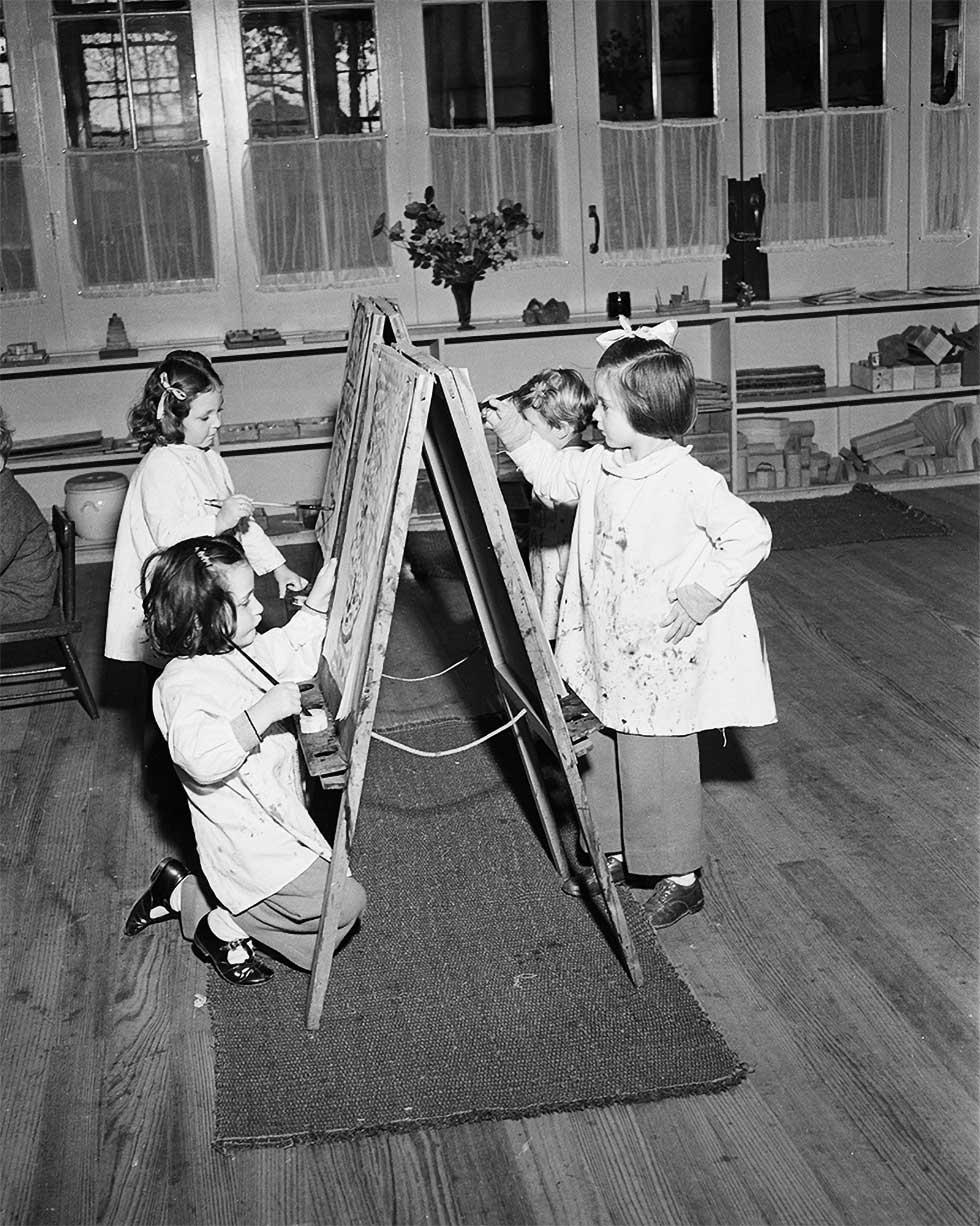 Four small children wearing smocks standing before painters easels painting.