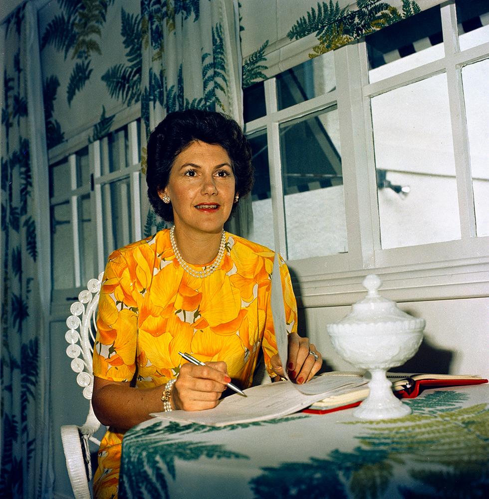Tammy Fraser seated at a table, pen in hand.  Green fern frond print on table cloth and curtains. 