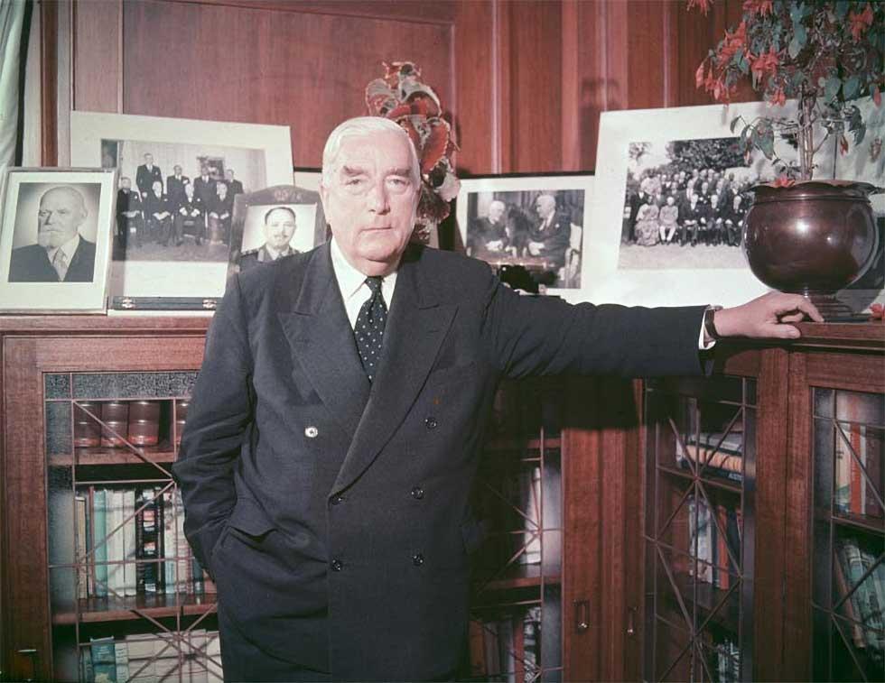 Robert Menzies standing in a wood-panelled room in front of a leadlight glass-fronted corner bookcase