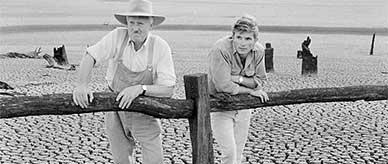 Two men leaning on a timber post and rail fence looking at the dry ground cracked into large blocks.