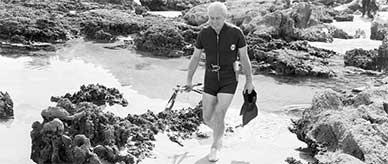 Prime minister Harold Holt walking by rock pools wearing a spring wet suit, carrying flippers, snorkelling gear and a speargun.