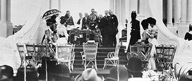 Men and women in formal Victorian attire gathered around a desk in the decorated Federation Pavilion, Centennial Park.