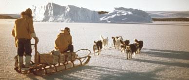 Seven huskies towing a dogsled carrying two people and their supplies across. 