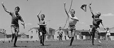Four children running toward the camera with kites while four children watch from behind.