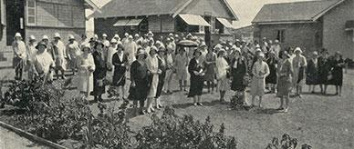 Women wearing hats – standing together on a lawn amongst large timber huts.