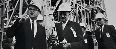 Men in suits wearing hard hats standing before a rocket gantry.