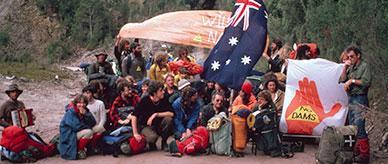 A group of protesters sitting on a dirt road holding flags and banners. 
