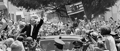 American president Lyndon B Johnson waving from his motorcade surrounded by a crowd.