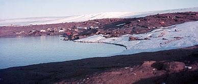 Mawson station in 1955. Buildings next to the sea, surrounded by snow and rock.