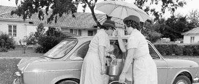 2 women, one holding an umbrella, place prepared meals into an Austin 1800 car.