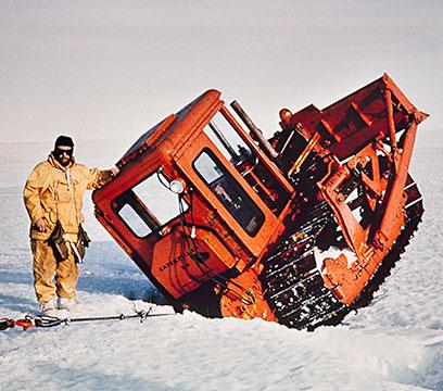 Man in yellow leaning against orange grader wedged in ice crevice.