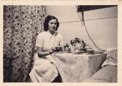 Eva Makay seated drinking tea at a table in front of a floral curtain. 