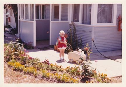 Trixie pushing a toy pram outside a blue house. 