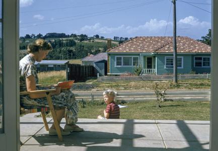 A woman sits reading a book on the front porch of her house. Her small son sits on the steps. 
